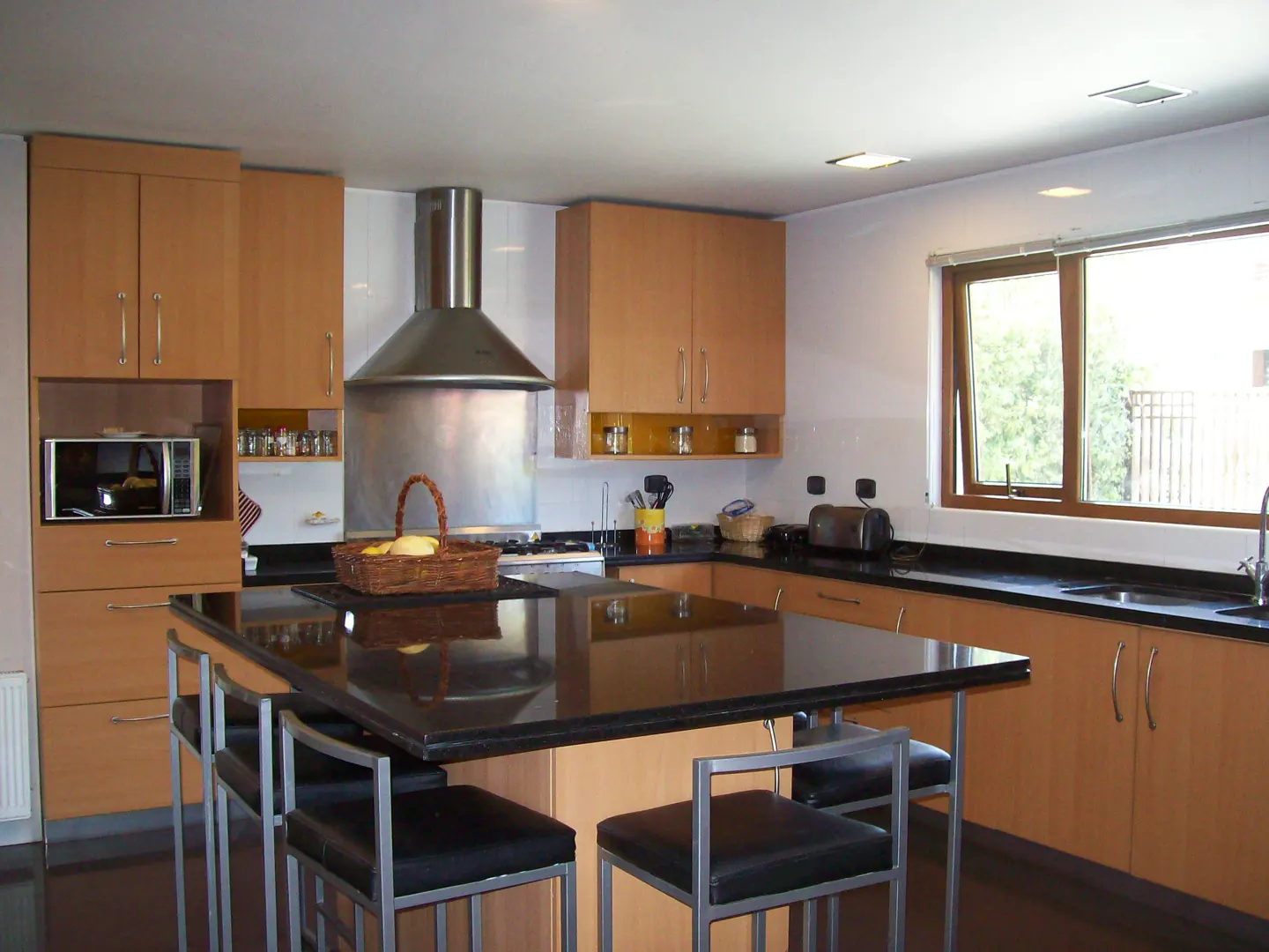 A kitchen with light wood cabinets, black countertops, and a stainless steel range hood. A black island with bar stools is in the foreground.