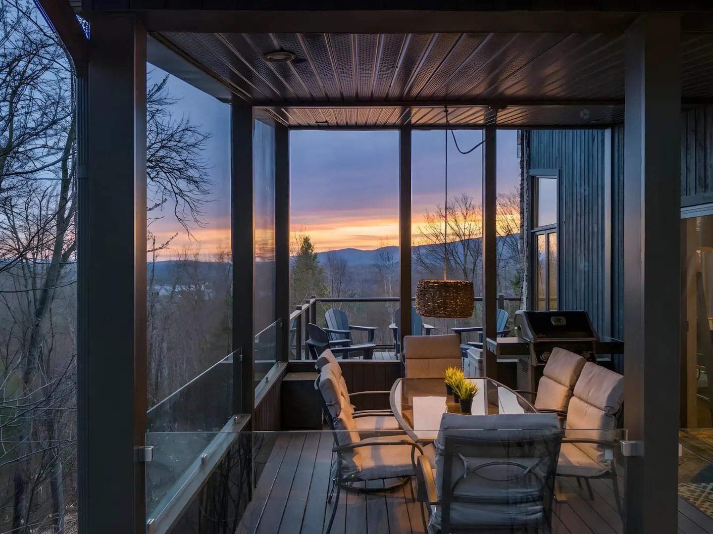 A screened-in porch with a dining table and chairs overlooks a mountain range at sunset.