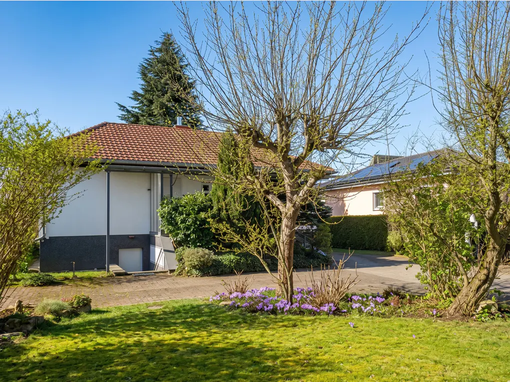 A single-story house with a red tile roof, white walls, and a dark gray foundation, viewed from a green lawn with trees and purple flowers.