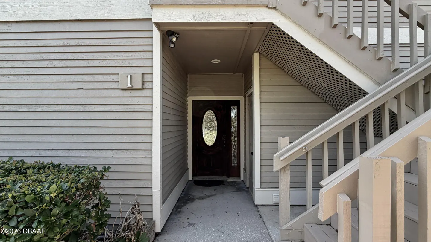 Exterior view of a condo unit with light gray siding, a dark wood door with oval glass, and stairs leading to an upper level.