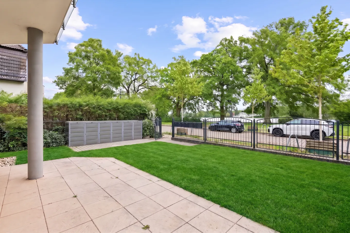 A patio with beige tiles leads to a green lawn, a fence, and two parked cars under a blue sky with trees.