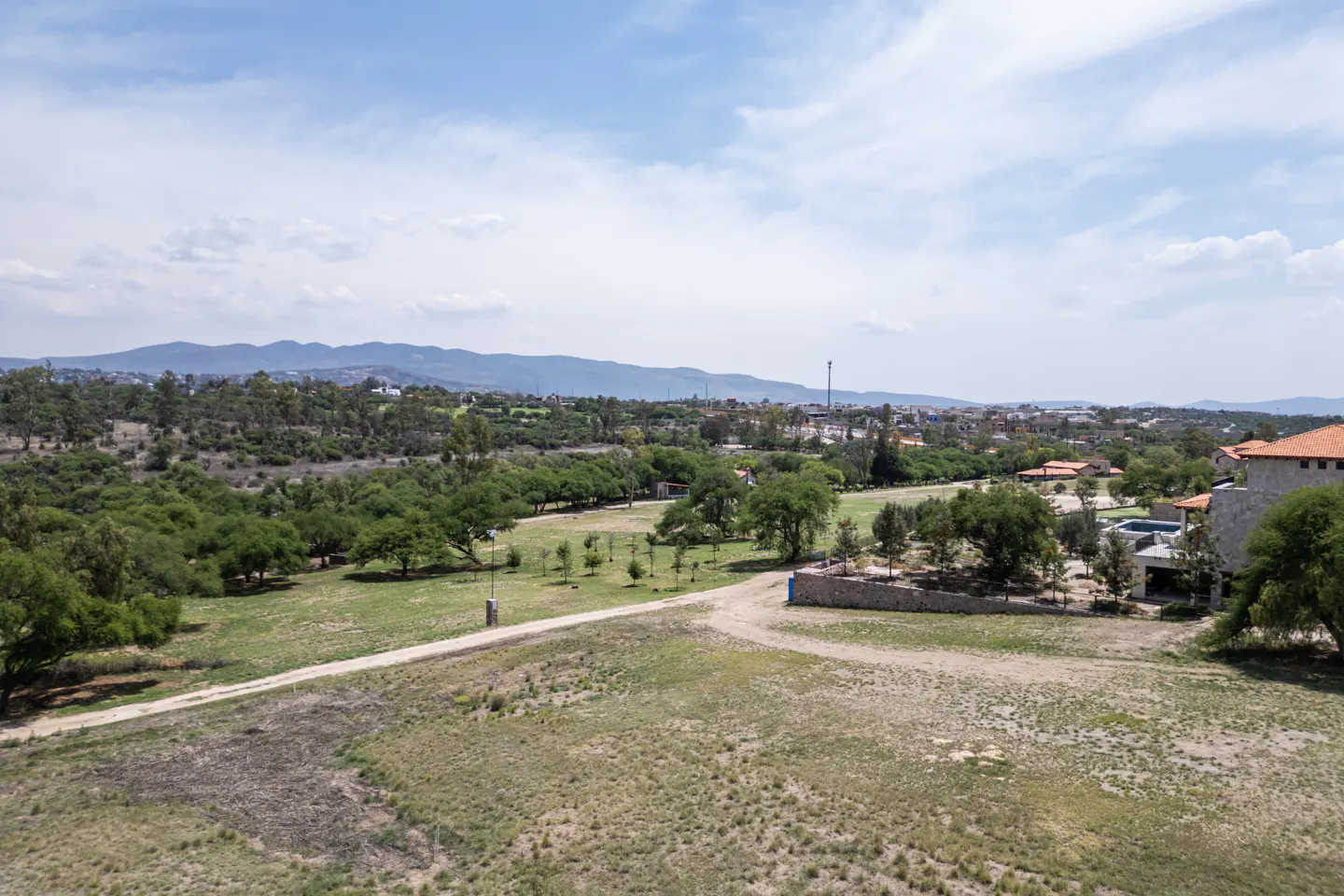 An aerial view of a grassy field with trees, a path, and mountains in the background under a blue sky with white clouds.