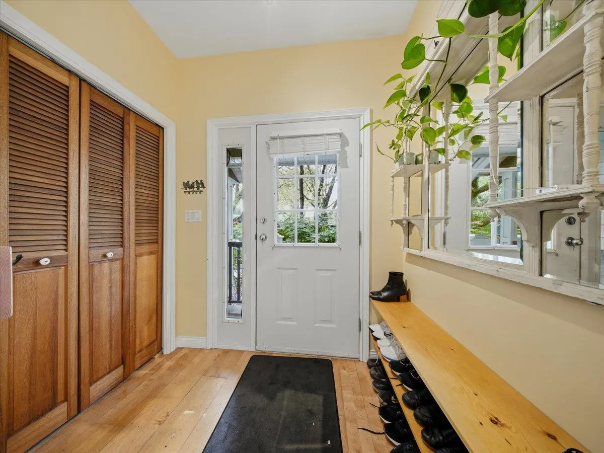 Entryway with wood floors, yellow walls, and a white door. A wood bench holds shoes under a decorative mirror with plants. A closet is on the left.
