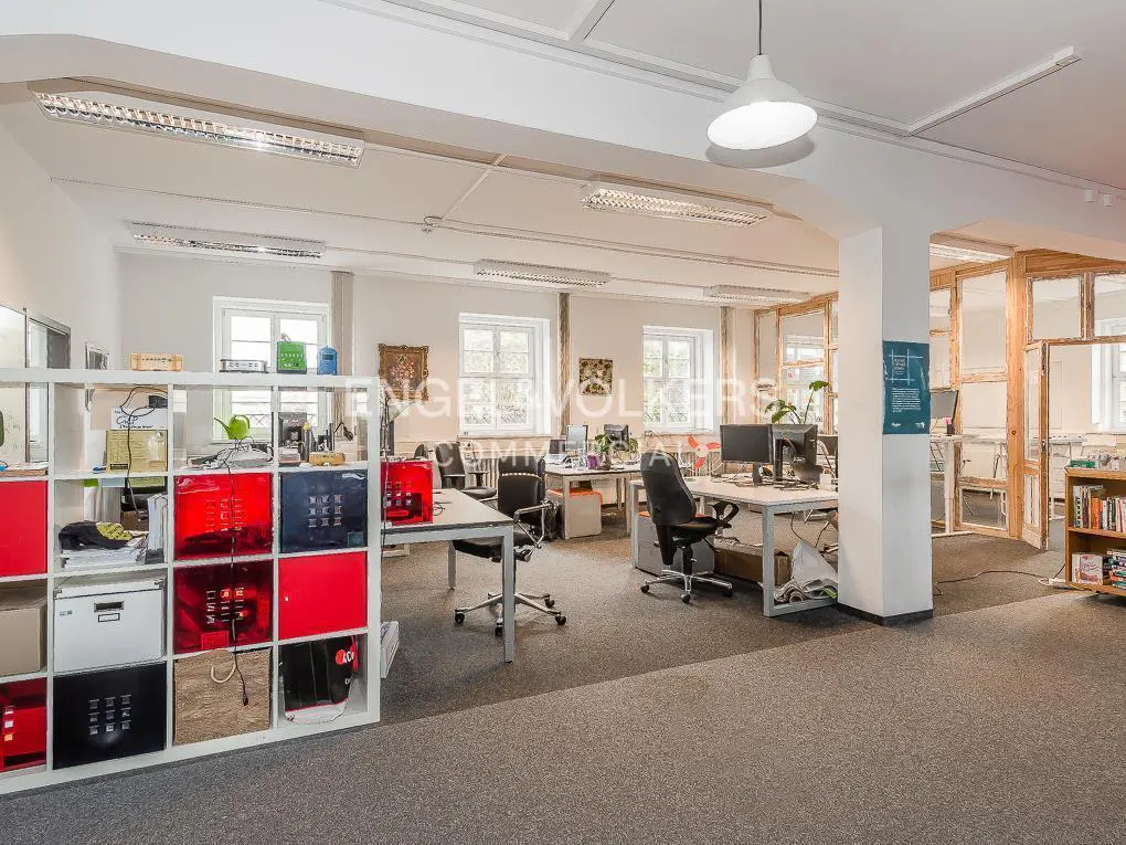 Bright, open office space with desks, chairs, and storage. White walls, gray carpet, and natural light from windows. Modern lighting fixtures.