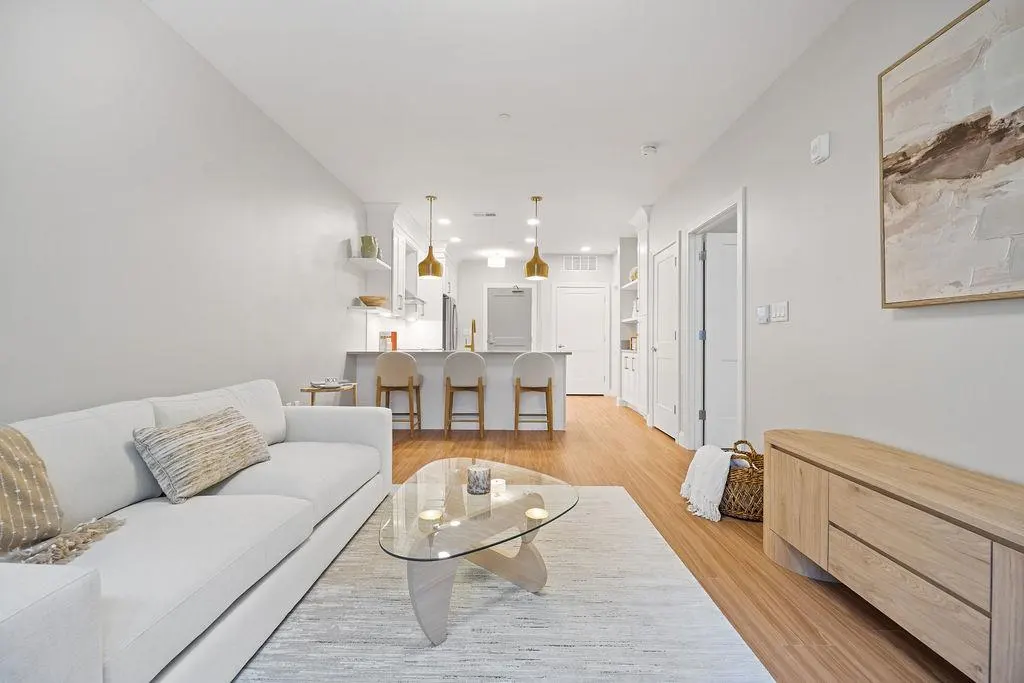 Bright, modern apartment living room with white sofa, glass table, and open kitchen. Artwork hangs on the wall.