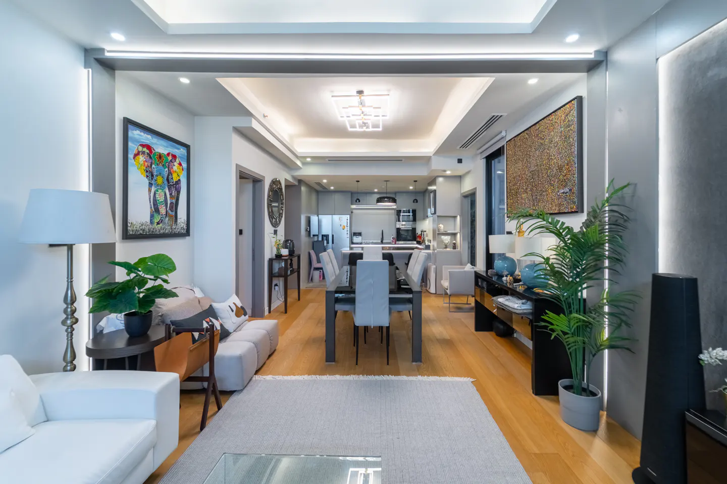 A modern apartment interior with a living room, dining area, and kitchen. The color palette is neutral with pops of color from artwork.