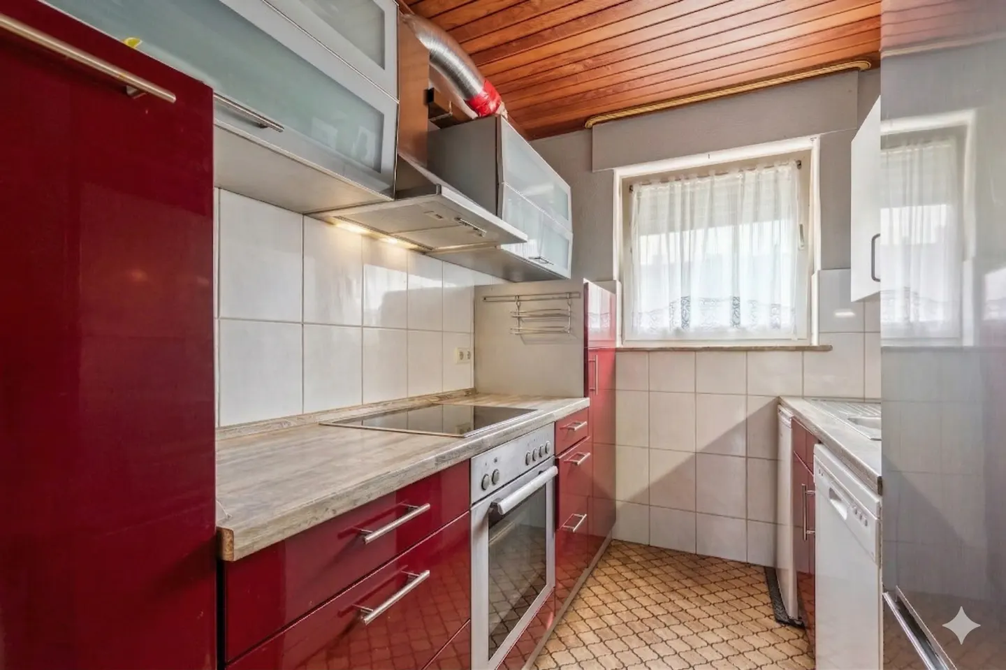 A kitchen with red cabinets, white tile backsplash, and a wood ceiling. A window with white curtains is visible.