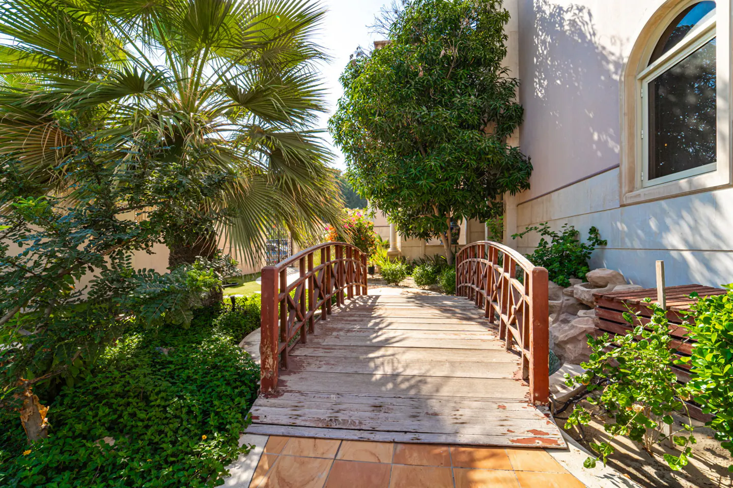 Wooden bridge with red railings over a small creek, surrounded by lush greenery and a palm tree, leading to a house.