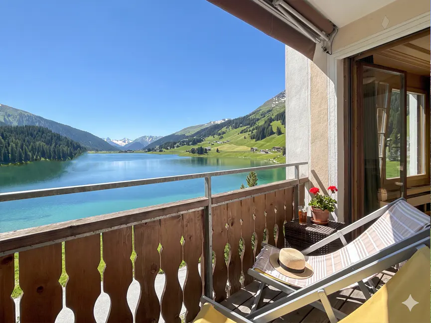 Balcony view of a turquoise lake and mountains. A lounge chair, hat, and drink sit on the balcony.