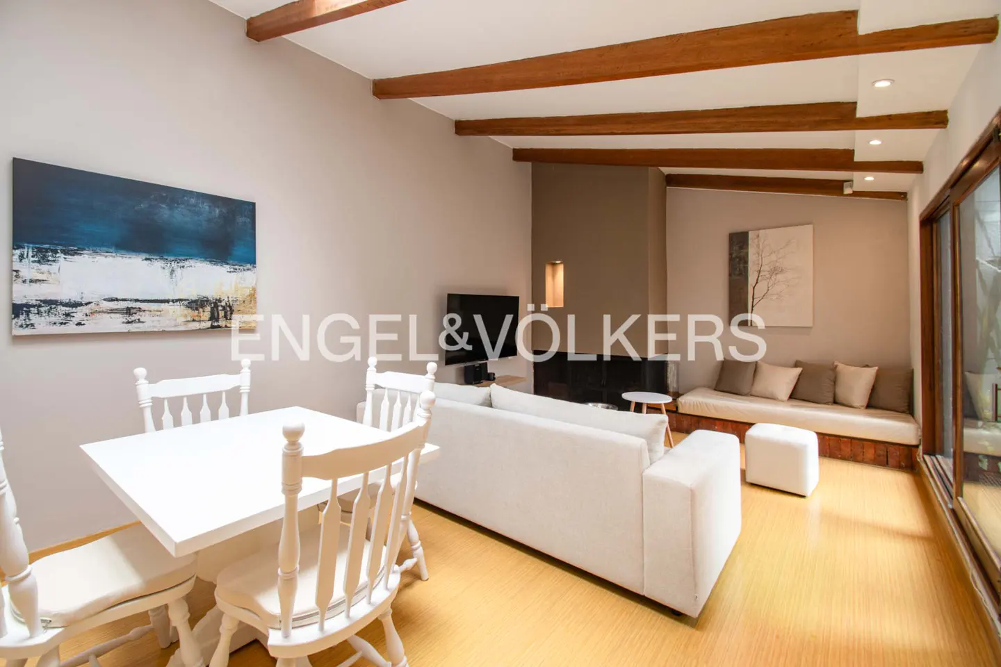 Living room with white furniture, wooden beams, and art. A white table and chairs are in the foreground. A white sofa and daybed are in the background.