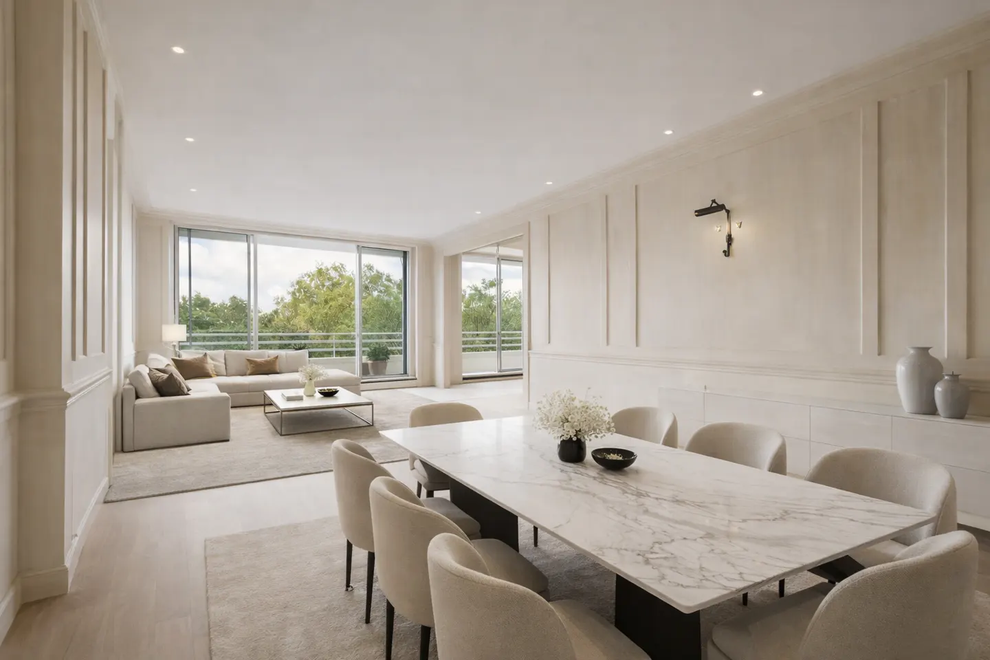 Bright, open-plan living and dining area with a marble table, beige chairs, and a white sofa near a large window overlooking greenery.