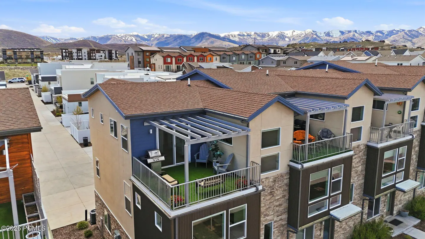 Aerial view of modern townhouses with brown roofs and balconies, set against a backdrop of snow-capped mountains under a blue sky.