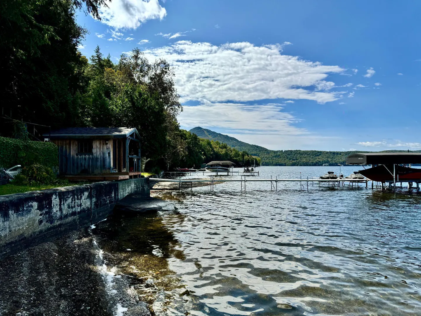 Lake view with a small wooden cabin on the shore, a dock extending into the water, and mountains in the background under a blue sky with clouds.