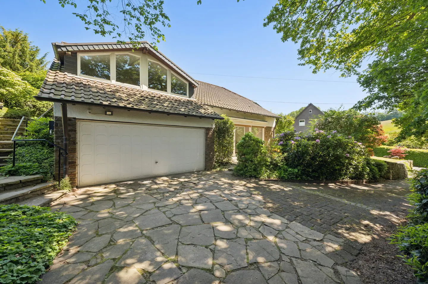 A house with a white garage door and stone driveway, surrounded by lush green trees and bushes under a clear blue sky.