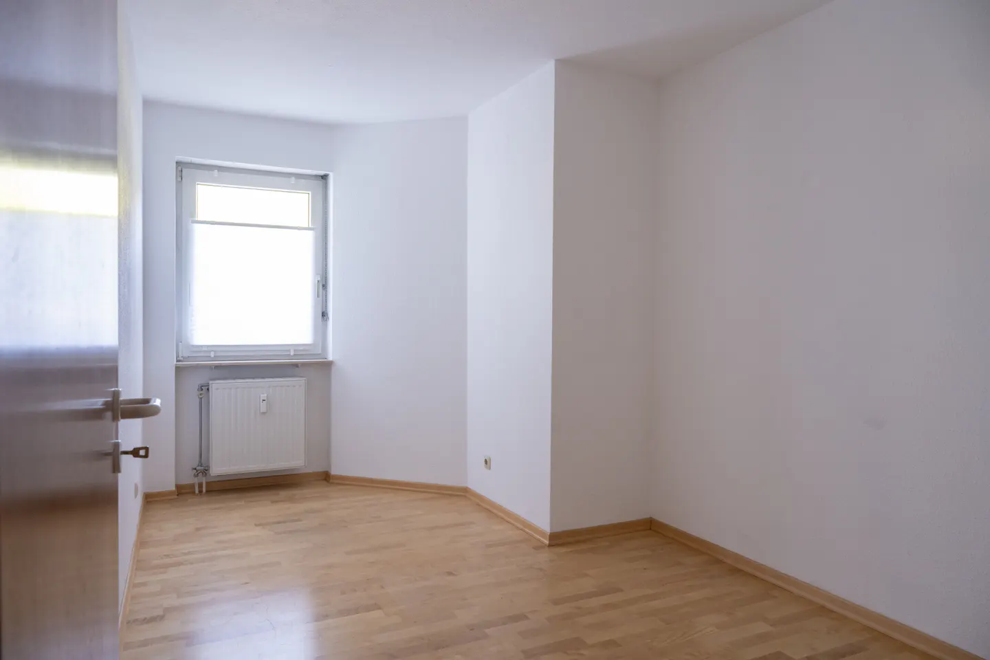 Bright, empty room with white walls and light wood floor. A window with blinds and a radiator are visible. A silver door is partially open.