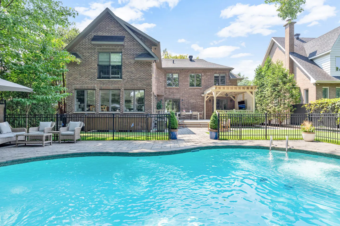 Backyard view of a brick house with a pool, patio furniture, and a pergola. The pool is turquoise and surrounded by a stone patio.