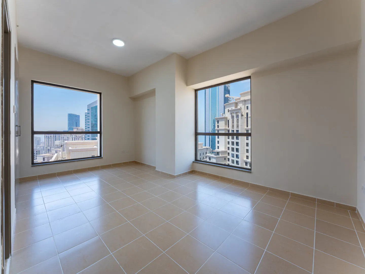 Empty room with beige walls and tile floor. Two windows show city views.