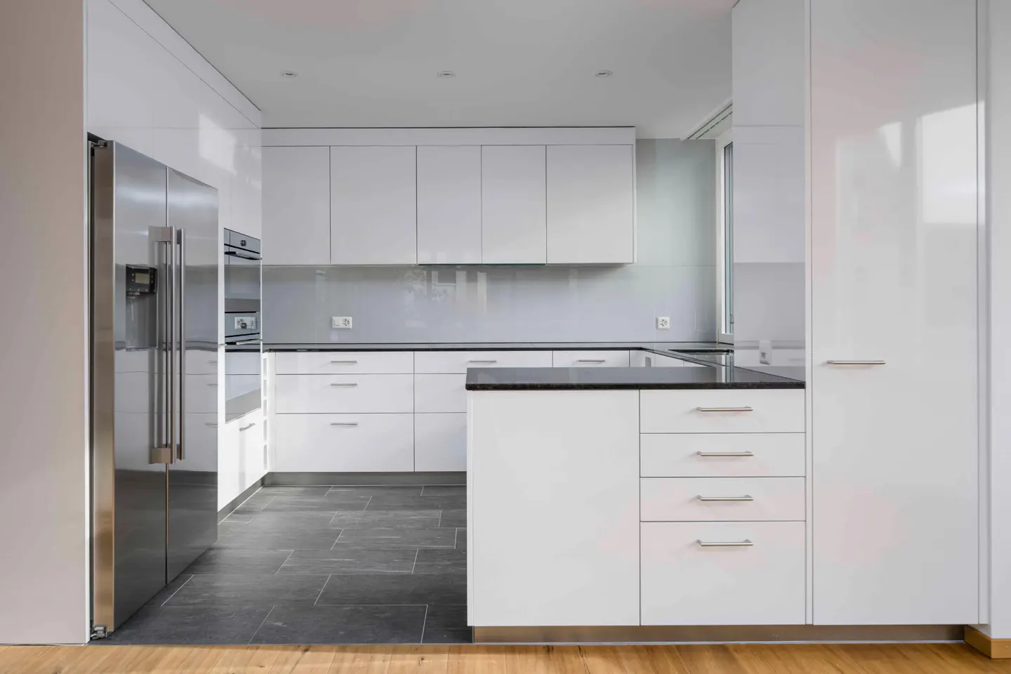 Modern kitchen with white cabinets, black countertops, and stainless steel refrigerator. Gray tile floor and light wood flooring.