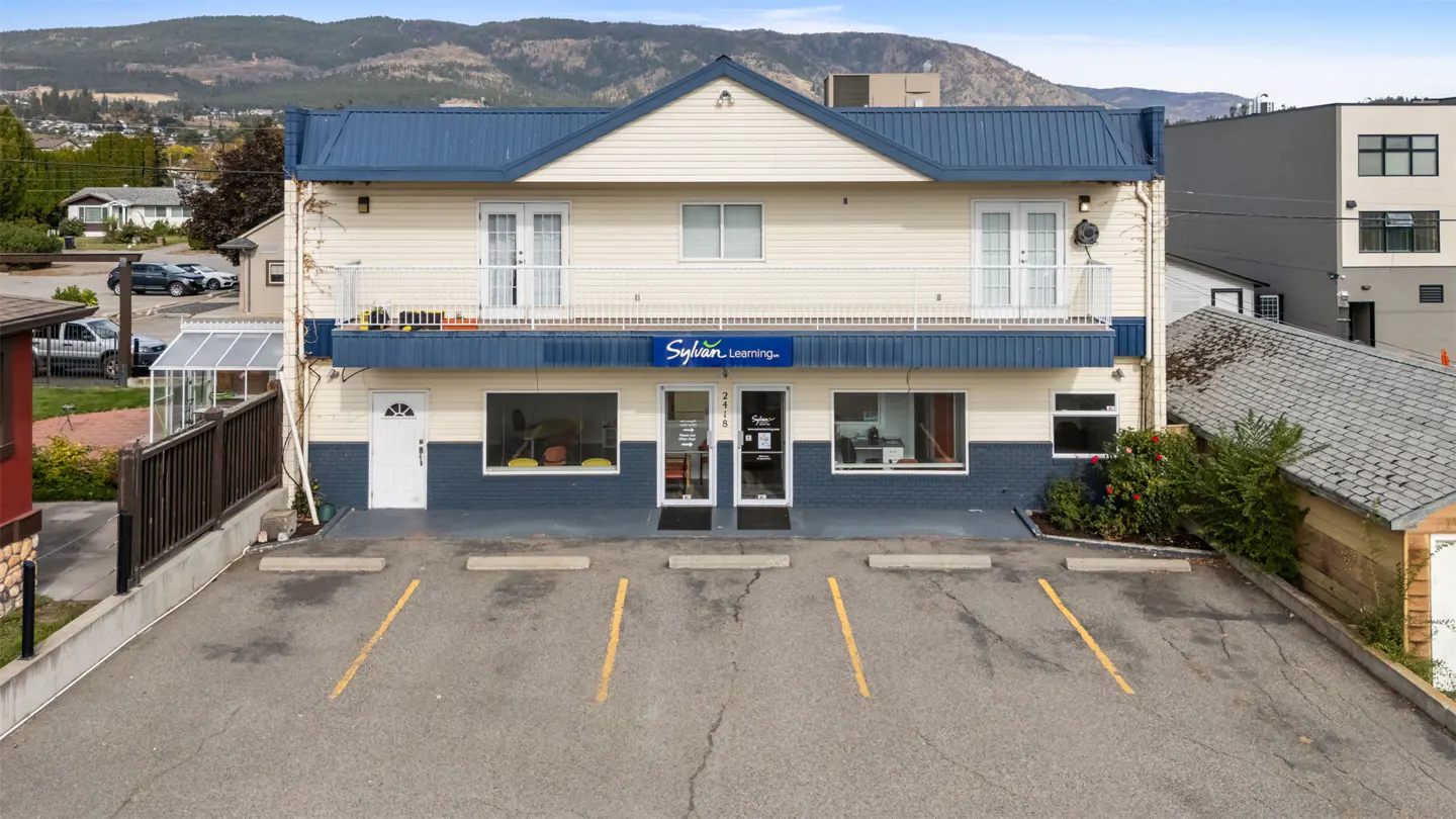 Two-story building with blue roof and "Sylvan Learning" sign. Parking lot with yellow lines in front. Mountains in background.