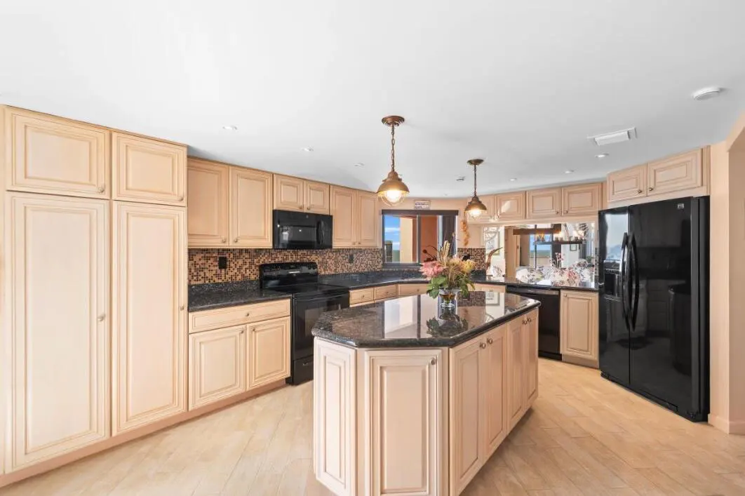 A kitchen with light wood cabinets, black appliances, and a granite island with flowers.