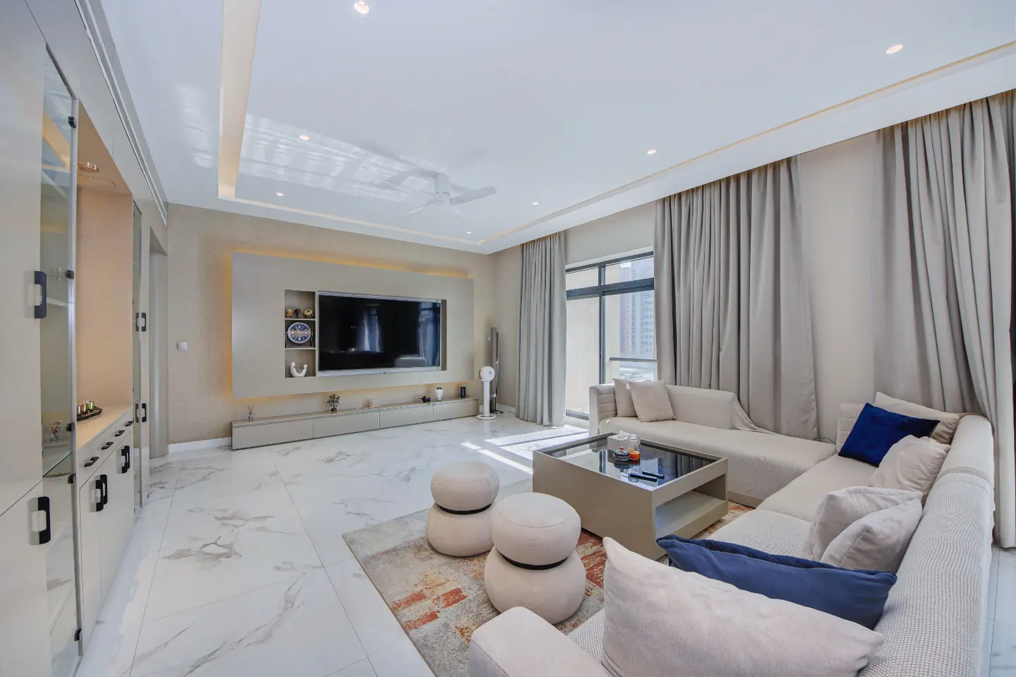 Bright living room with white marble floors, a large TV, and a beige sectional sofa with blue pillows.