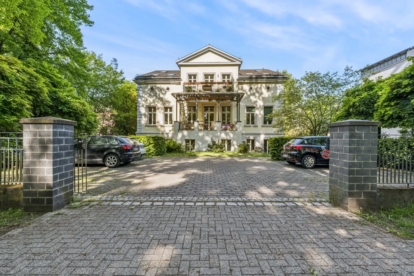 A grand, light-colored villa with a balcony, viewed from a paved driveway flanked by brick pillars and parked cars.