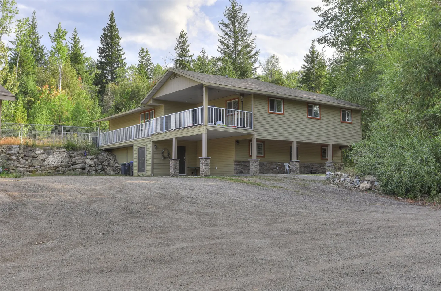 Two-story, light-green house with a white-railed balcony, supported by stone pillars, surrounded by trees and a gravel driveway.