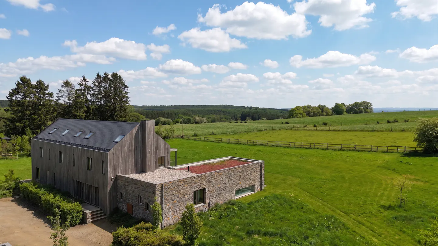 Modern home with wood and stone exterior, black roof, and green roof extension, set in a green field under a blue sky.