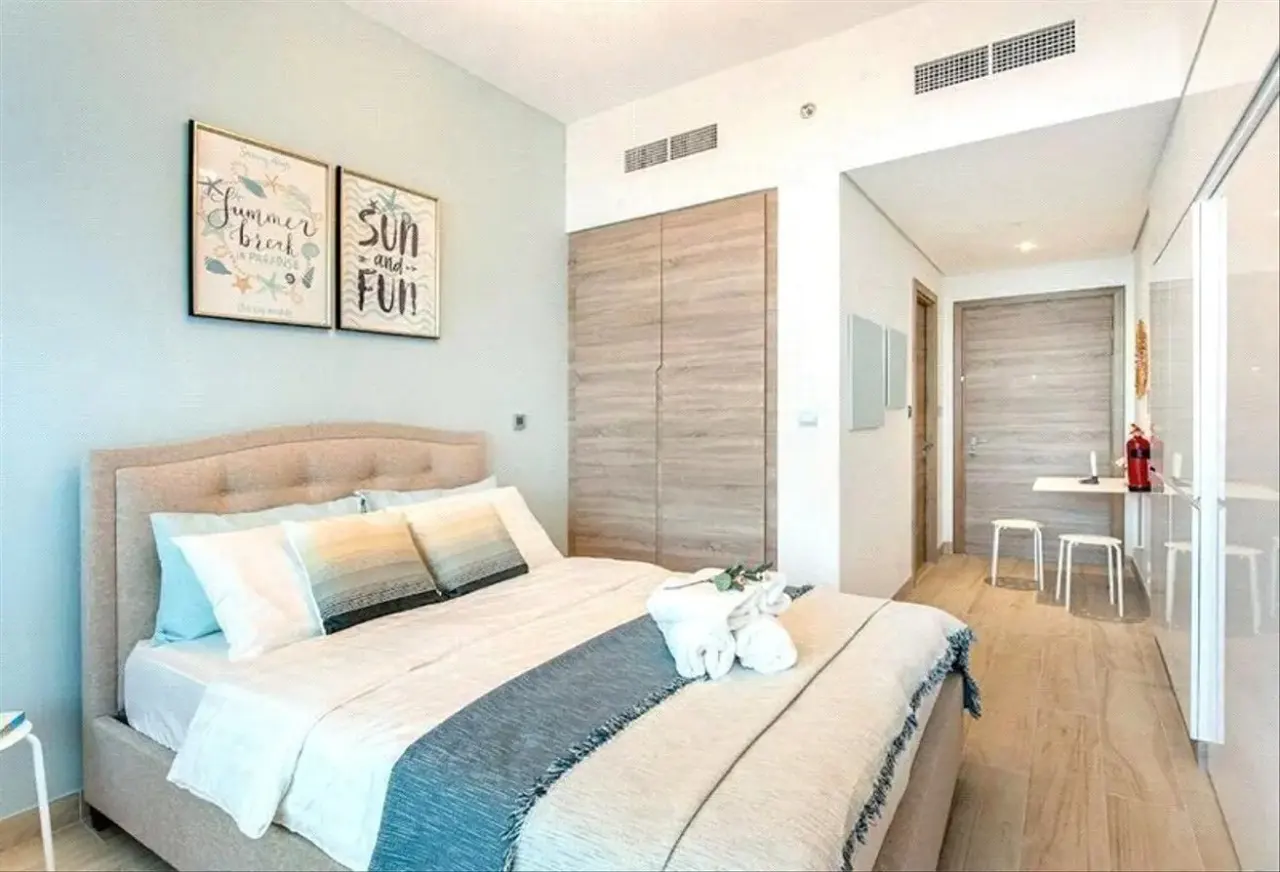 Bedroom with a beige bed, white linens, and blue accents. Two framed prints hang above the bed. A small table and chairs are visible in the background.