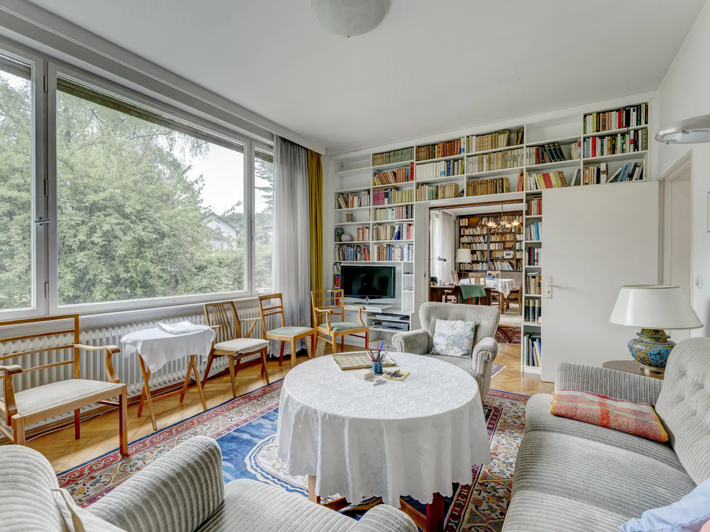 Living room with a round table, grey sofas, large windows, and floor-to-ceiling bookshelves filled with books.