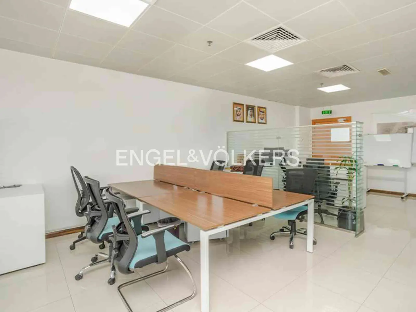Bright office space with white walls, tile floors, and a long wooden desk with blue chairs. Glass partitions and whiteboards are in the background.
