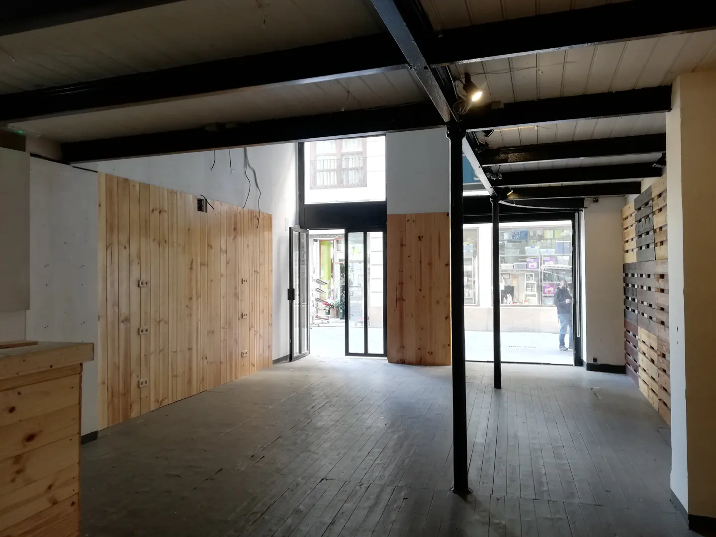 Empty retail space with wood floors, black metal beams, and wood paneled walls. Glass doors lead to a street view.