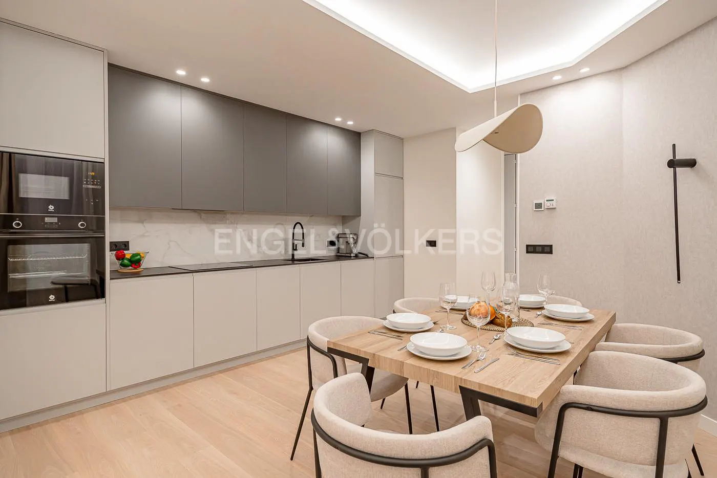 A modern kitchen and dining area with gray cabinets, a wooden table set for eight, and a unique pendant light.