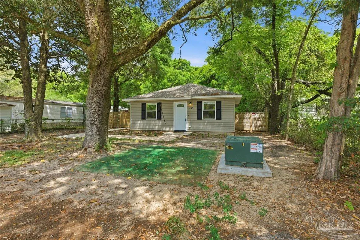 A small, one-story house with gray siding and black shutters, surrounded by trees and a sandy yard.