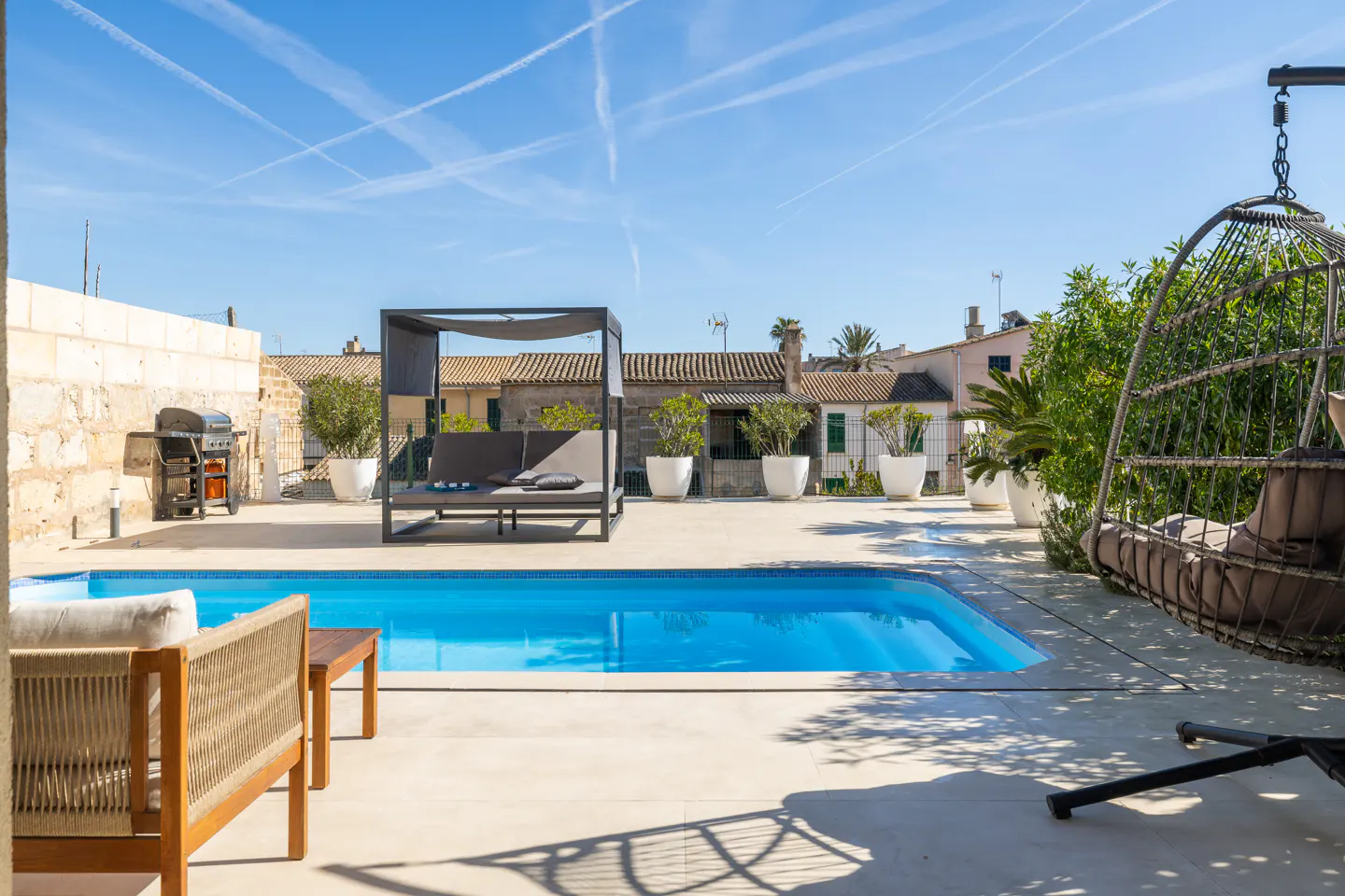Outdoor pool area with a blue pool, lounge chairs, a grill, and a hanging chair under a sunny blue sky.