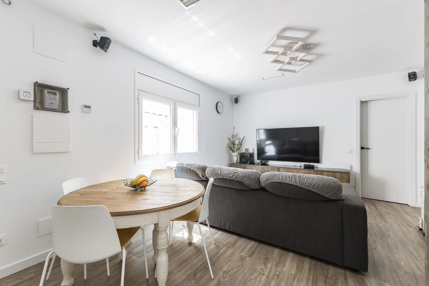Bright living room with white walls, wood floors, and a gray sofa. A round wooden table with white chairs sits near a window. A large TV is mounted on the wall.
