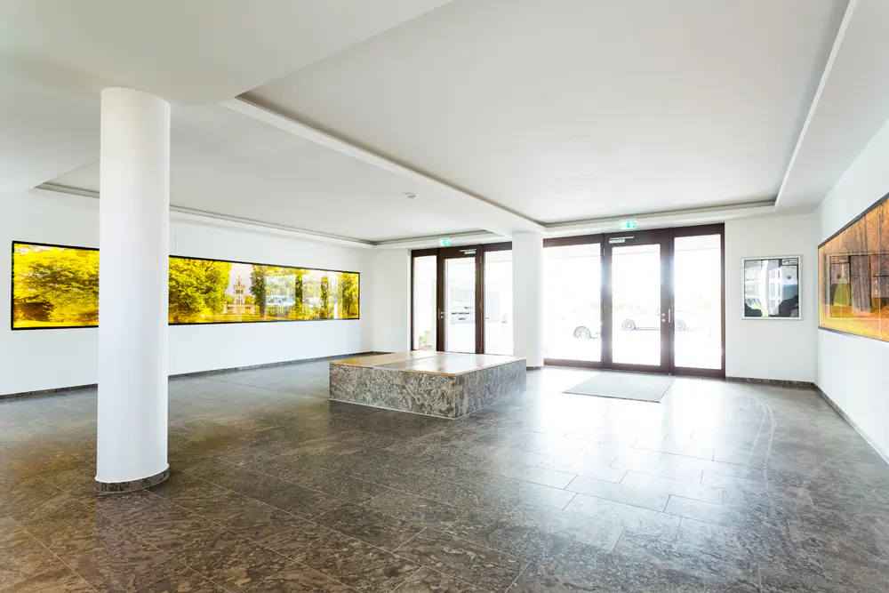 Bright lobby with marble floors, white walls, and a pillar. Large landscape art hangs on the walls. Double doors lead outside.