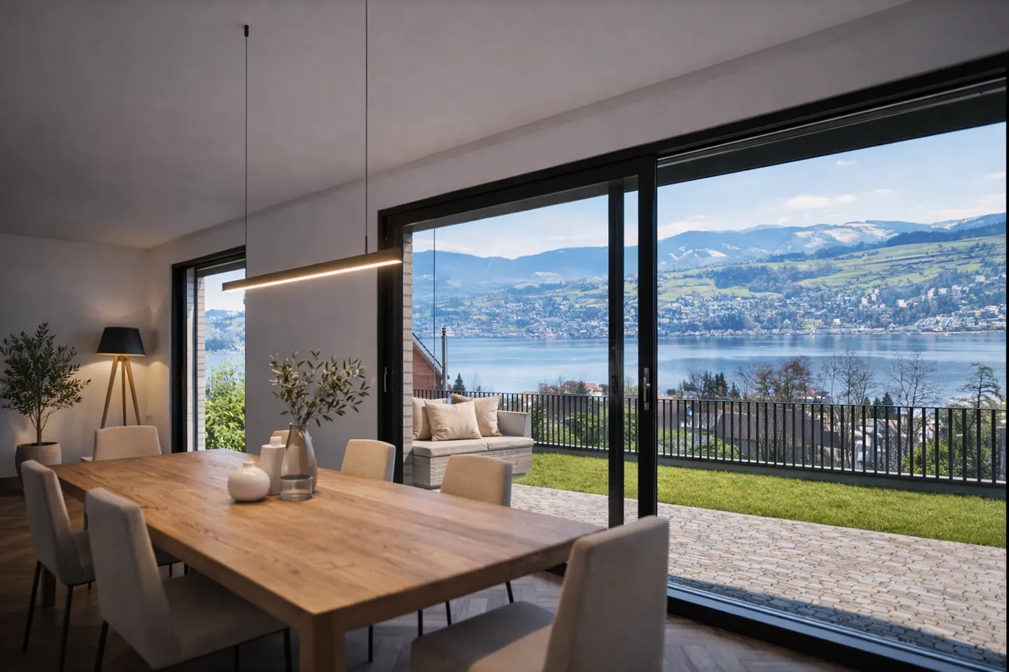 Bright dining room with a long wood table, beige chairs, and a view of a lake and mountains through sliding glass doors.