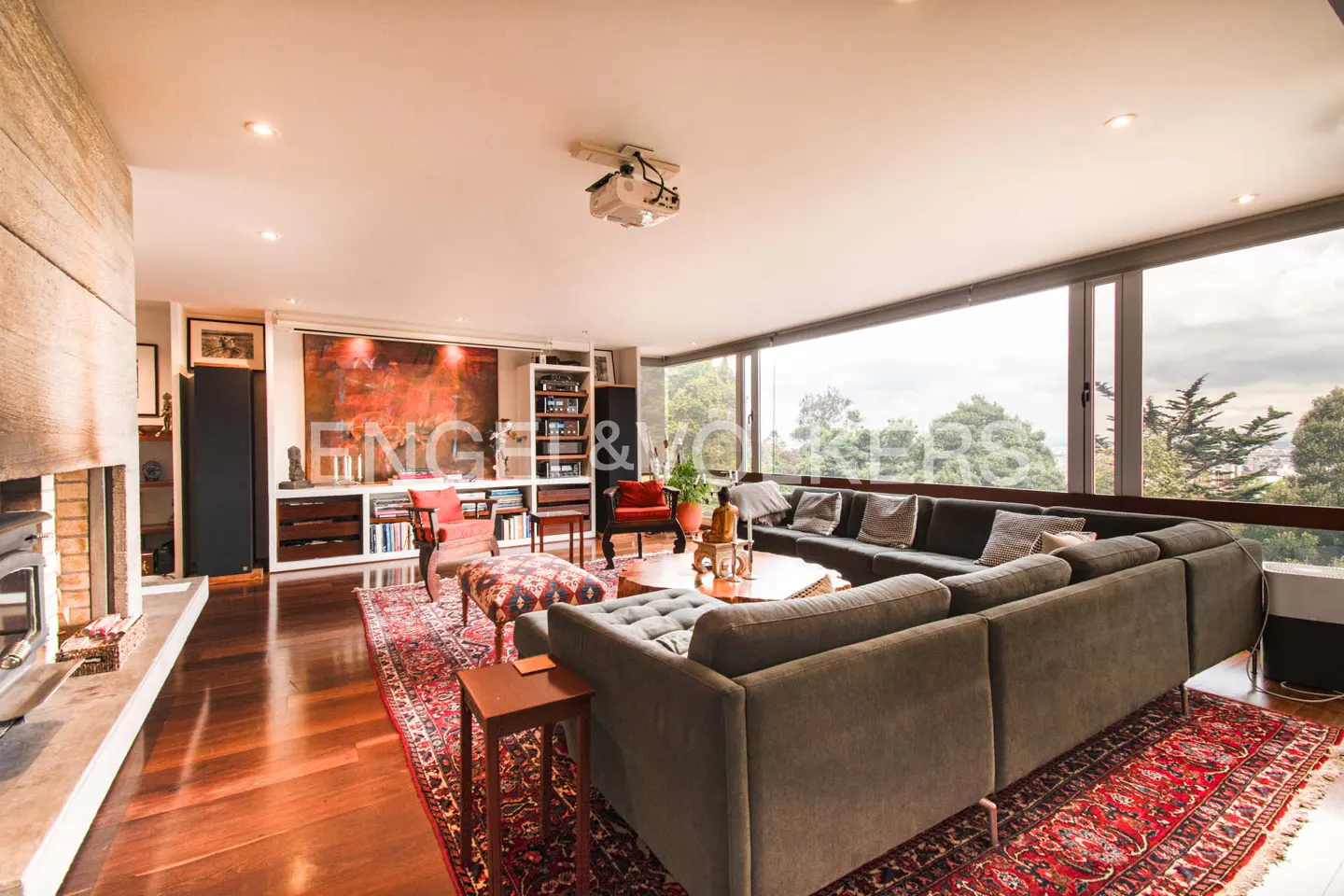 Living room with a large gray sectional sofa, red patterned rug, fireplace, and floor-to-ceiling windows with a view of trees.