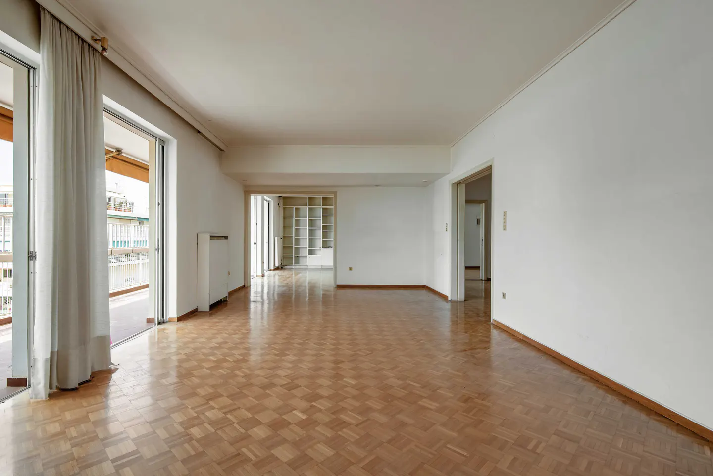 Bright, empty room with parquet floors, white walls, and balcony doors with sheer curtains. A built-in bookshelf is visible in the background.