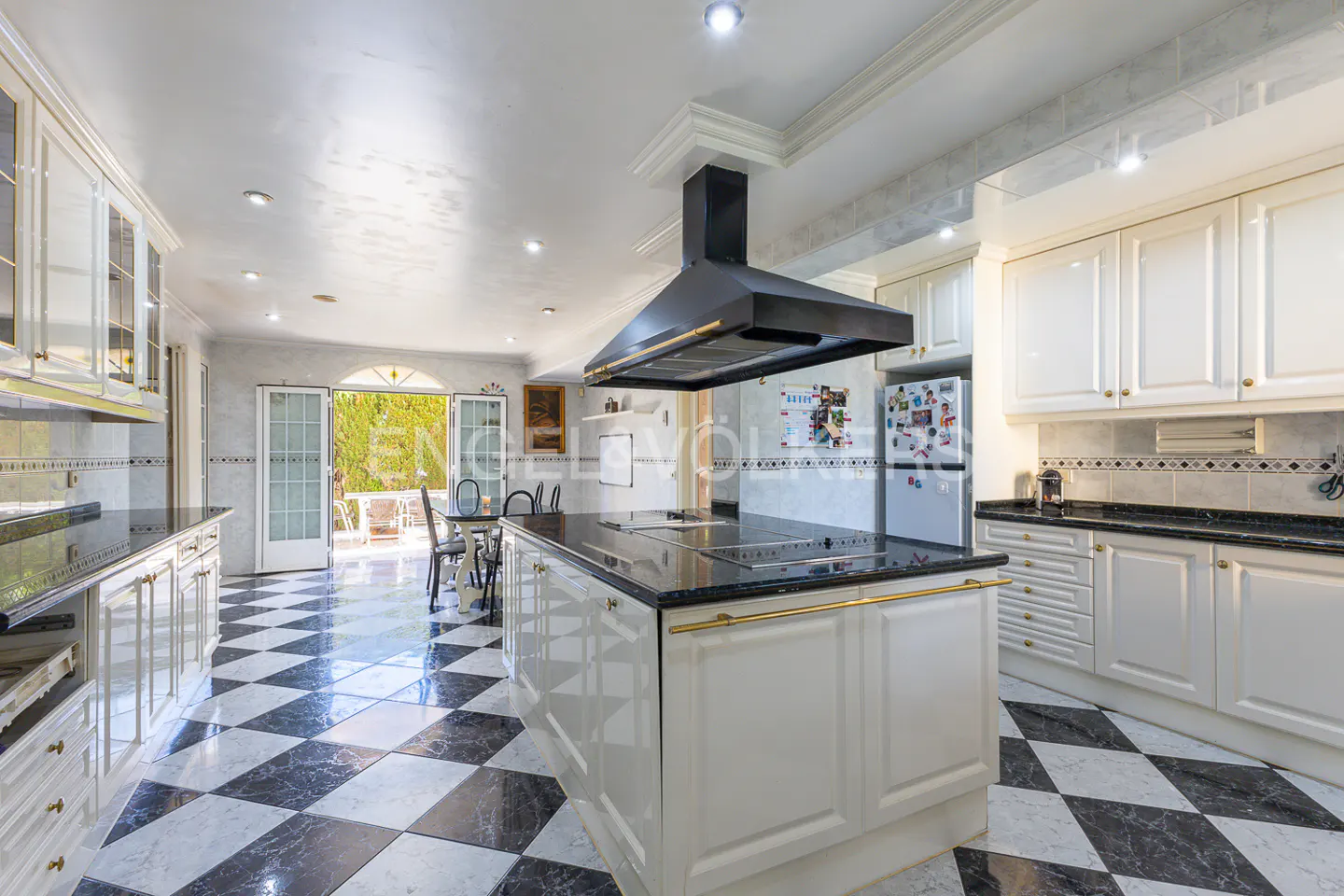 A bright kitchen with white cabinets, black countertops, and a black and white checkered floor. A black range hood hangs over the island. French doors lead to an outdoor patio.