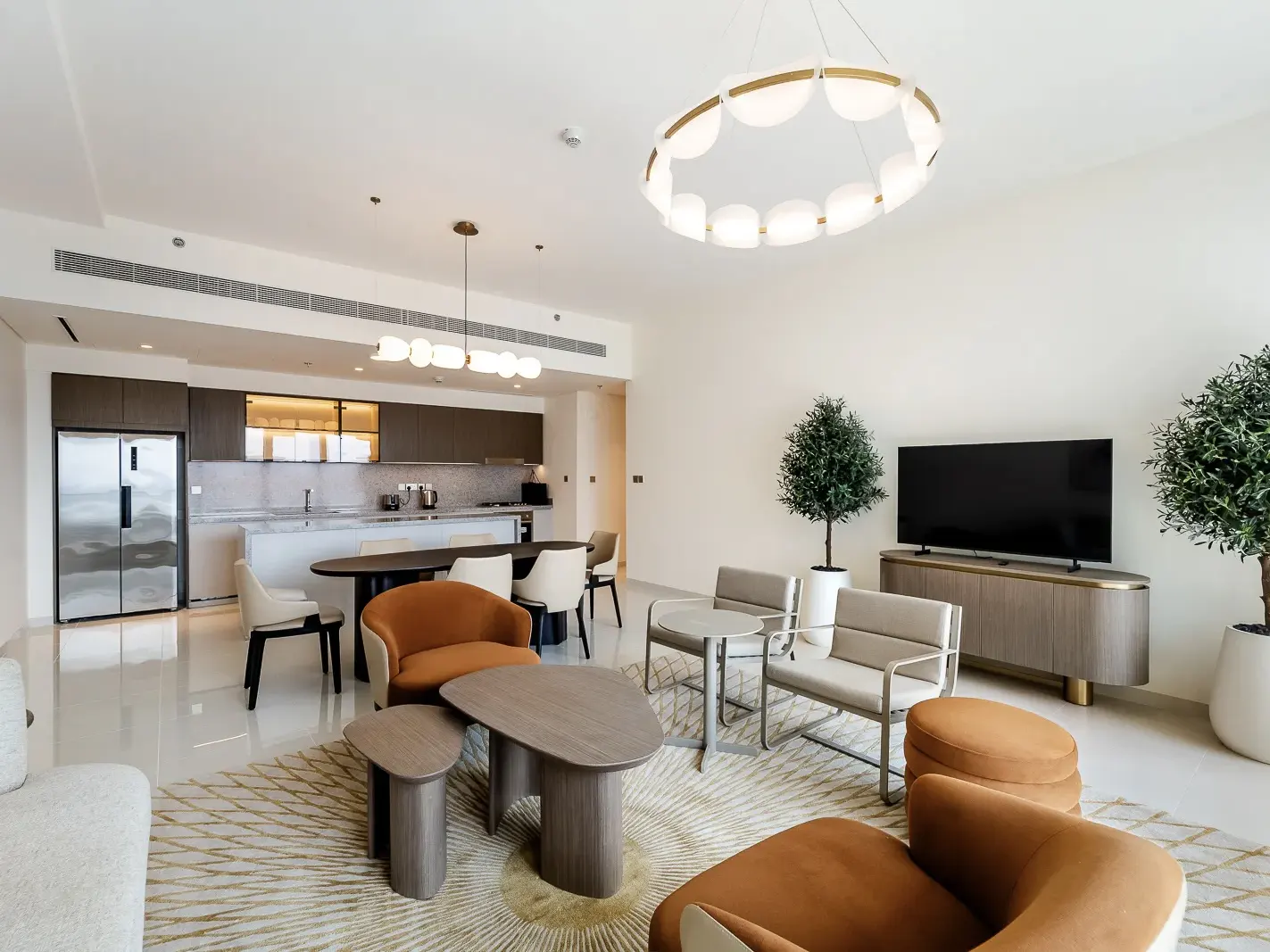 Bright, modern living room with kitchen, dining, and seating areas. Features brown cabinets, oval table, and orange chairs on a patterned rug.