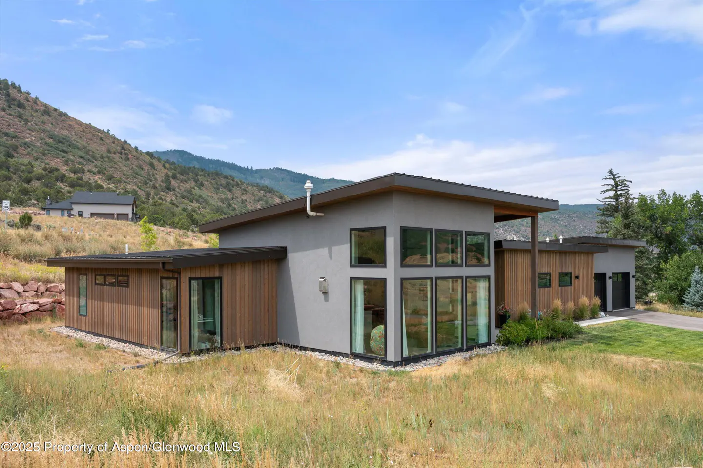 Modern home with gray stucco and wood siding, large windows, and a mountain backdrop under a blue sky.