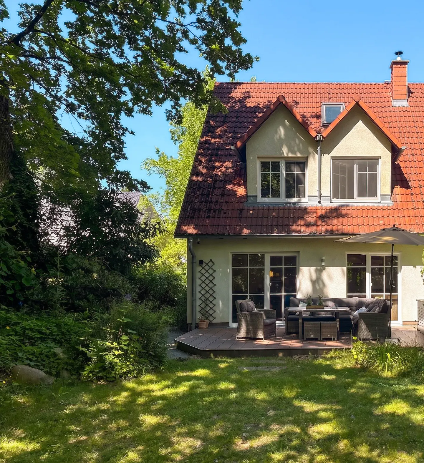 Backyard view of a two-story house with a red tile roof and a wooden deck with outdoor furniture. Green lawn and trees surround the house.