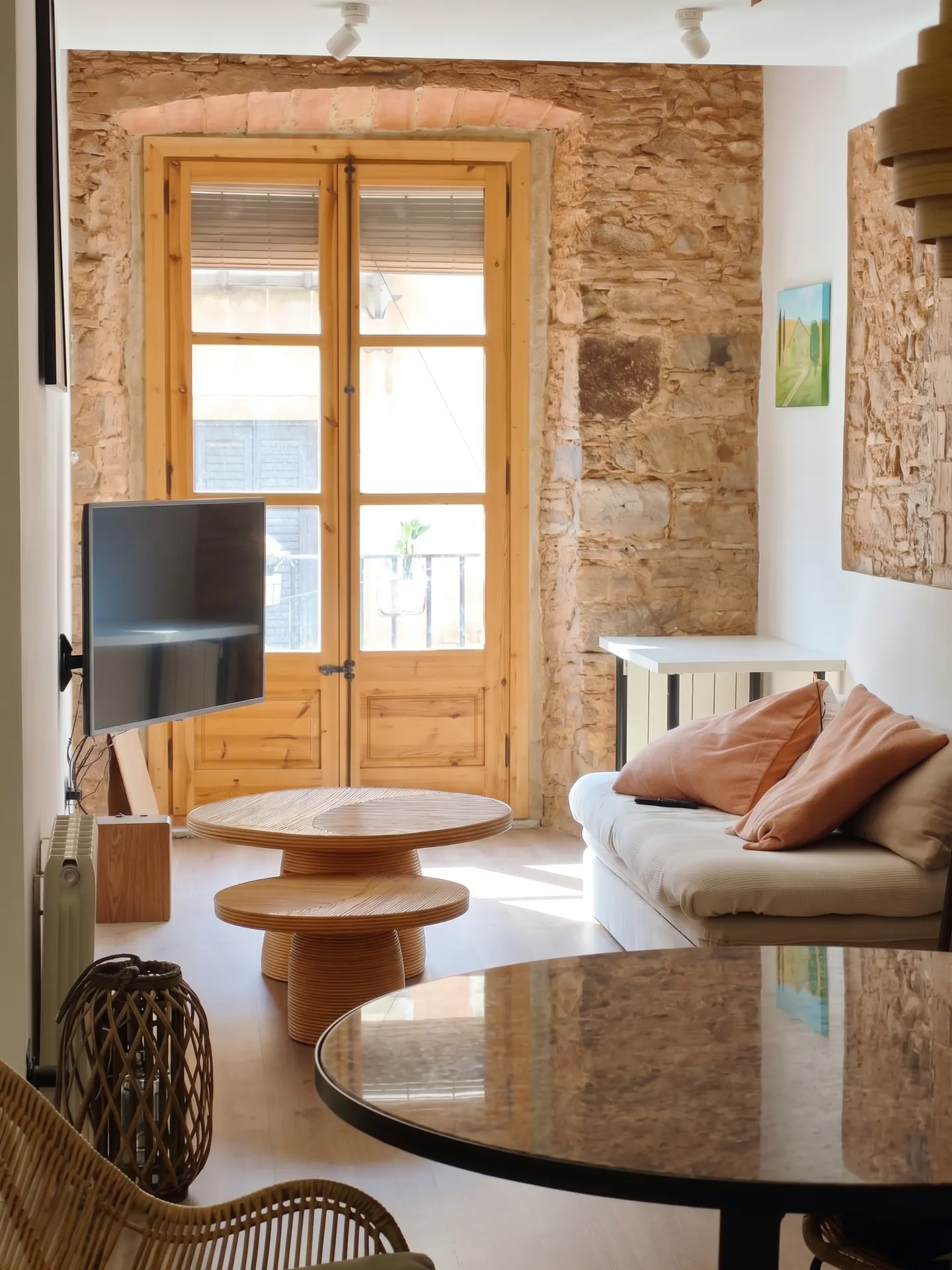 Living room with stone wall, wooden doors, and round tables. A white sofa with peach pillows sits near a painting. A TV is mounted on the wall.