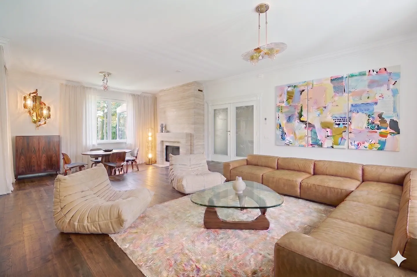 Bright living room with a brown leather sectional, glass coffee table, and colorful abstract art. A fireplace and dining area are in the background.