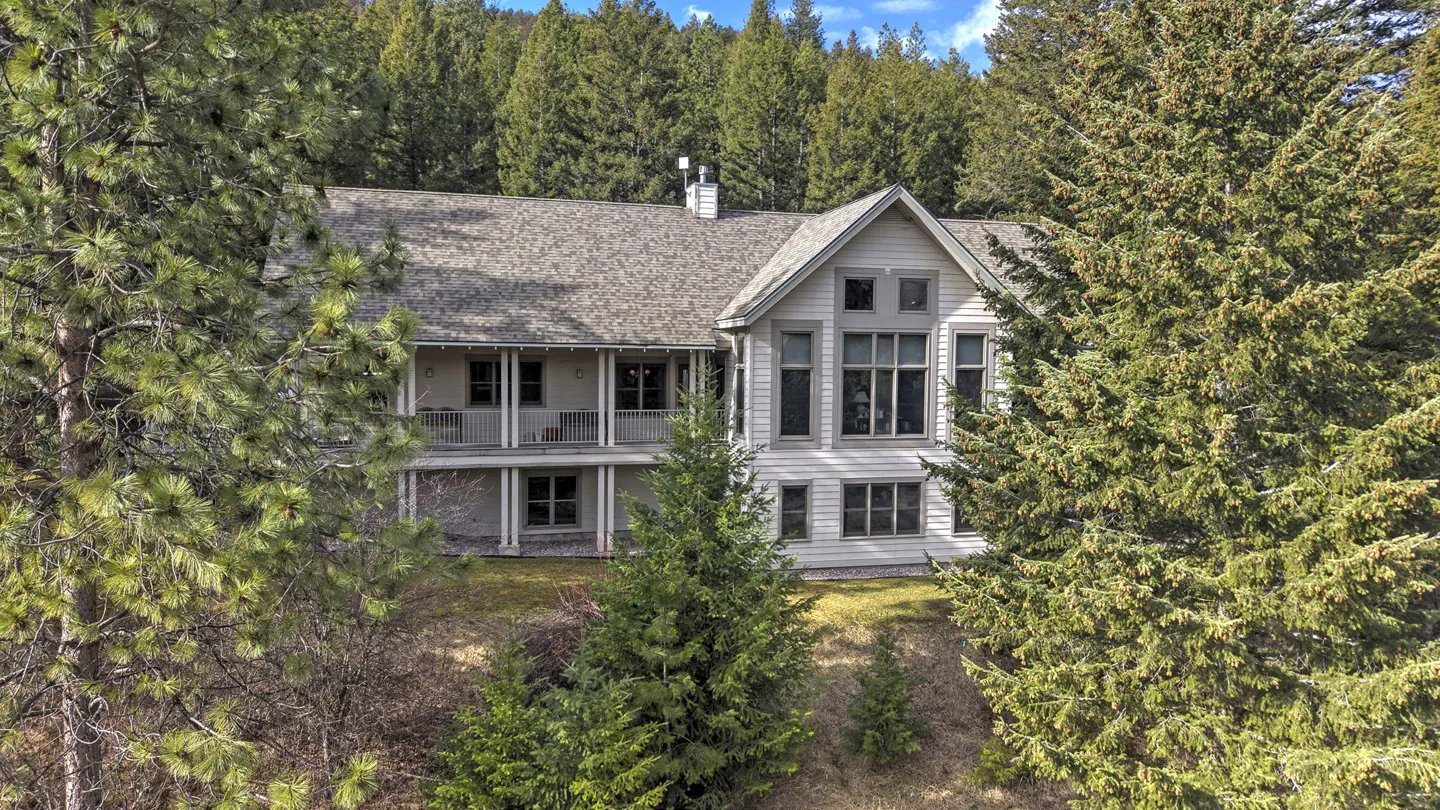 Two-story house with gray roof and siding, surrounded by green trees. Balcony on the second floor. Large windows.