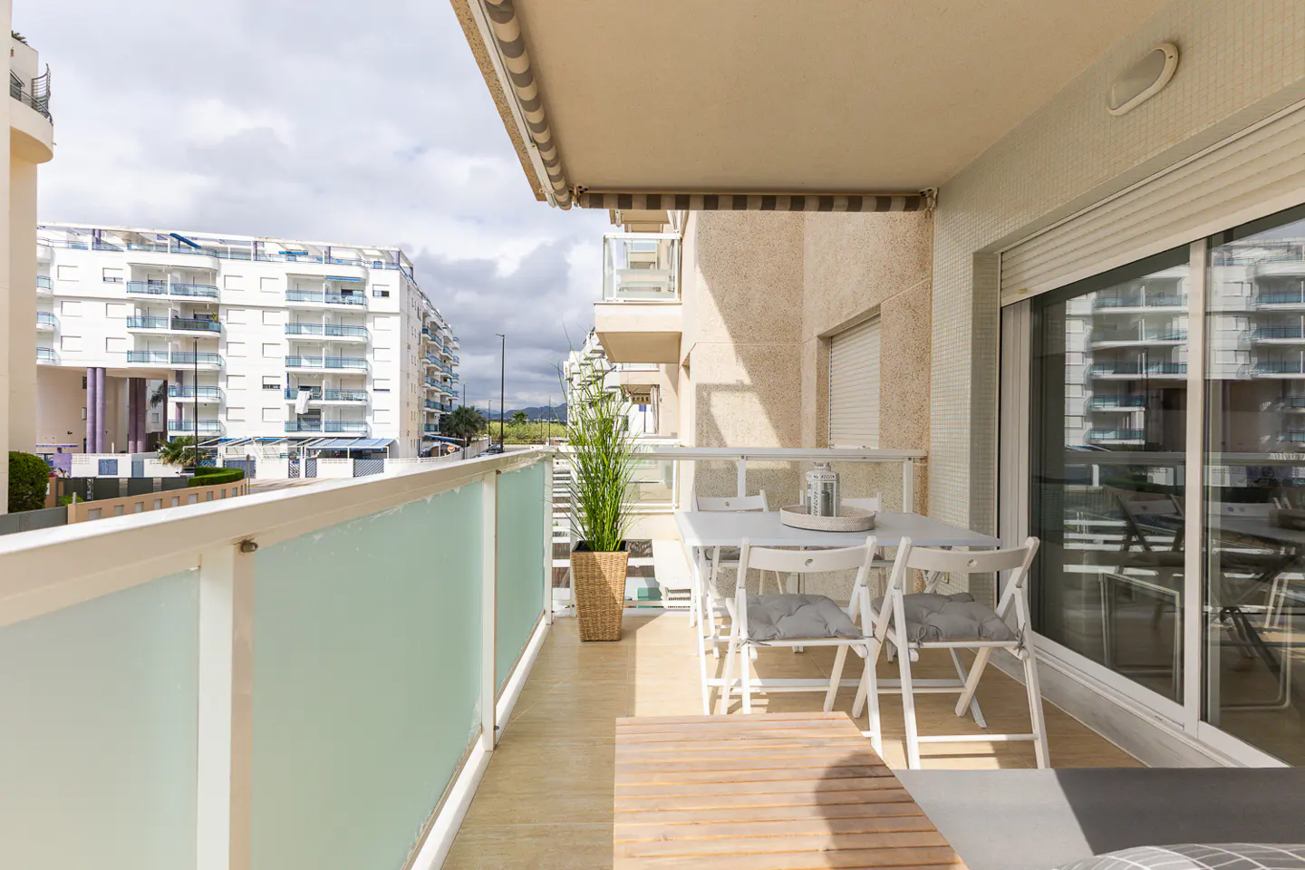 A balcony with a dining set, a potted plant, and a glass railing, overlooking a modern apartment complex under a cloudy sky.