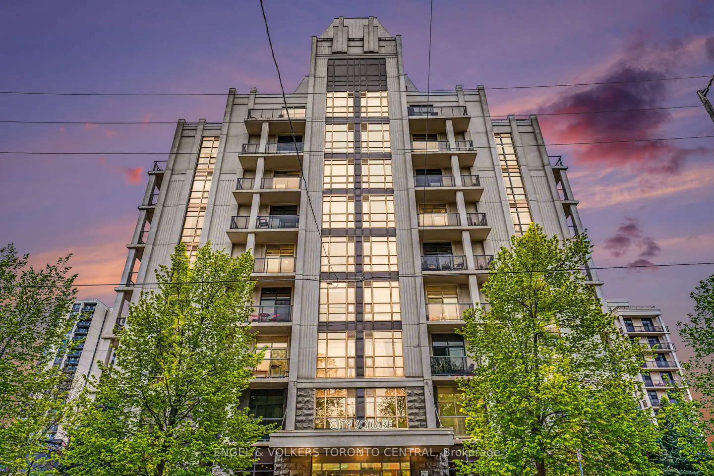 Exterior shot of a tall, light gray condo building with balconies, trees, and a colorful sunset sky.