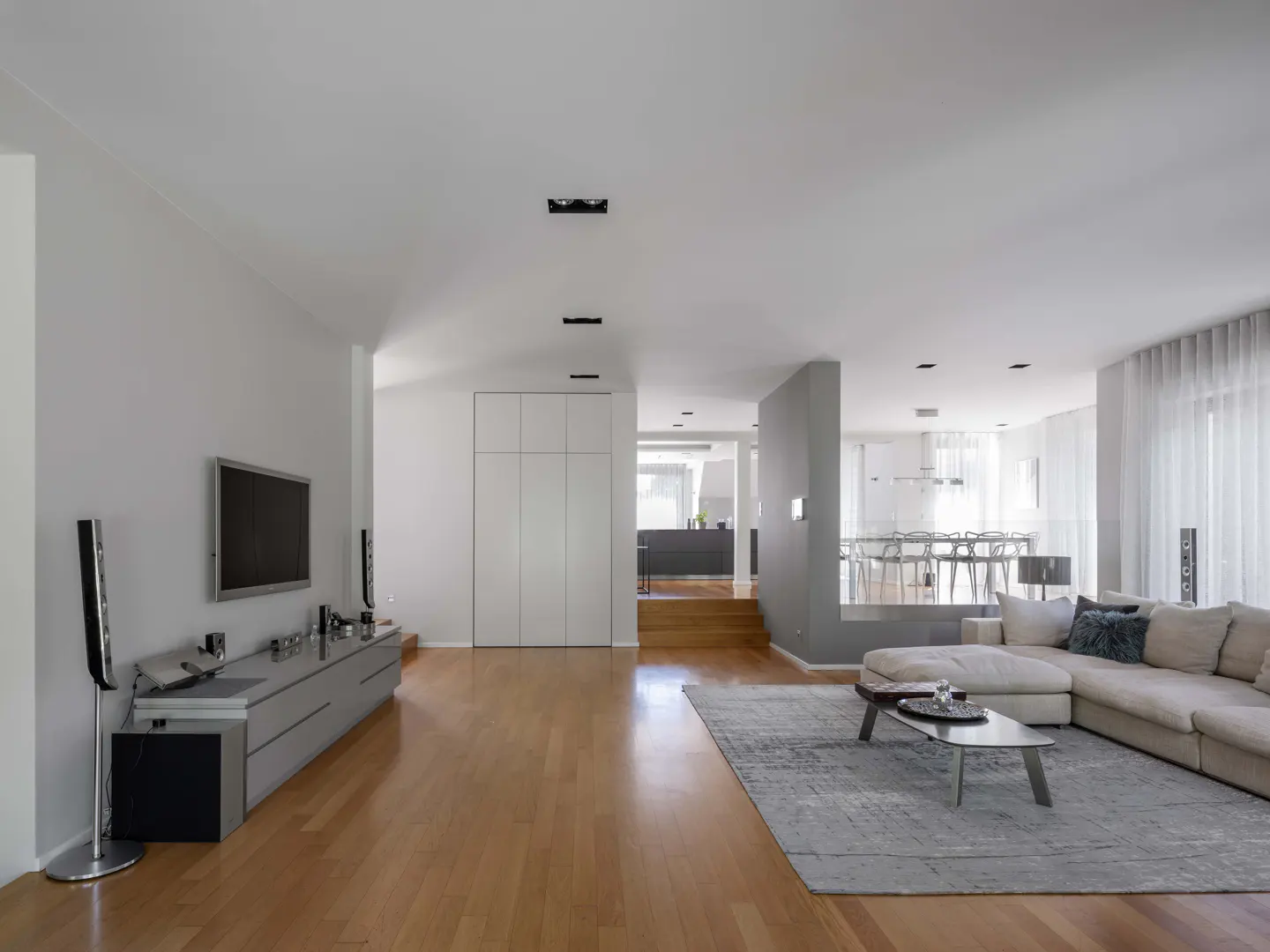 Bright, modern living room with hardwood floors, white walls, and a large sectional sofa. A TV and speakers sit on a gray console.